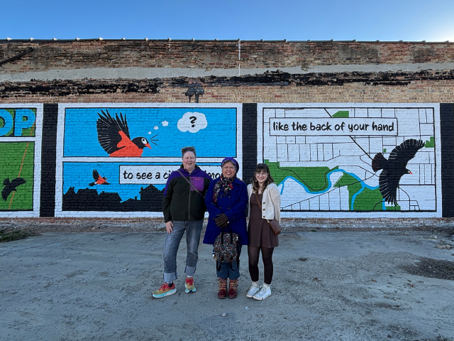 3 individuals stand in front of a colorful mural depicting birds flying above a map of Racine, Wisconsin