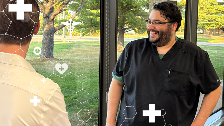 Photo of students standing by a bank of windows in Tallent Hall, in scrubs and a white coat