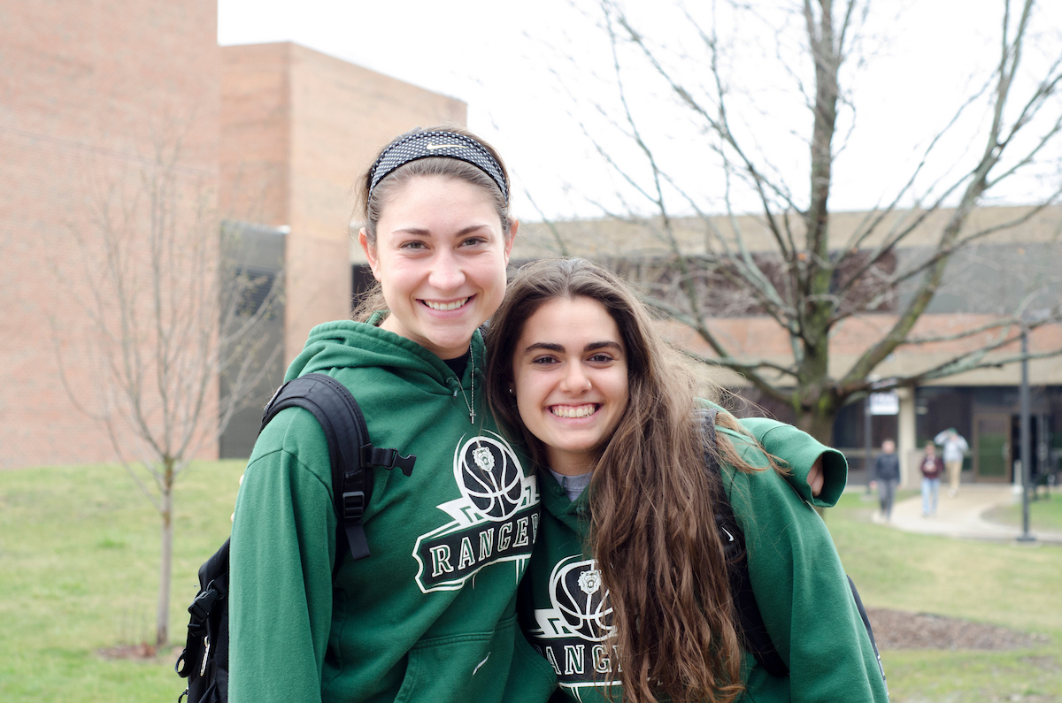 Two young women smiling and posing together outdoors on a campus, wearing matching green 