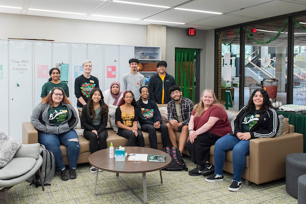 Gemini said A group of thirteen young adults poses in a bright lounge. Four individuals stand behind a long tan couch where nine others are seated.