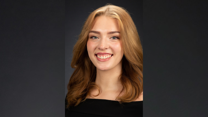 A headshot of a smiling young woman with long, wavy strawberry-blonde hair and a black off-the-shoulder top against a dark grey background.