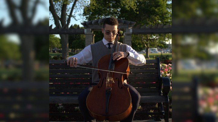 A person in formal attire playing the cello while seated on a park bench.