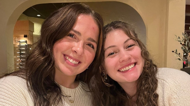  Gemini said Two young women with long, wavy hair smile closely together in a warm, indoor setting. They are both wearing white knit sweaters.