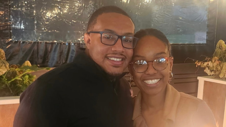 A smiling man and woman wearing glasses stand close together in a warmly lit indoor setting.