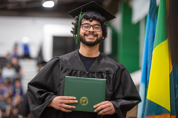 A smiling graduate in a black cap and gown holds a green diploma cover featuring a gold university seal.