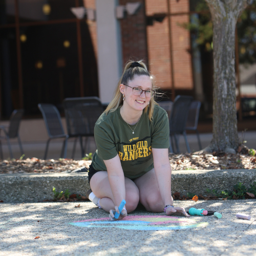 Girl outside wearing a Ranger Tshirt smiling as she's using chalk on the ground. 