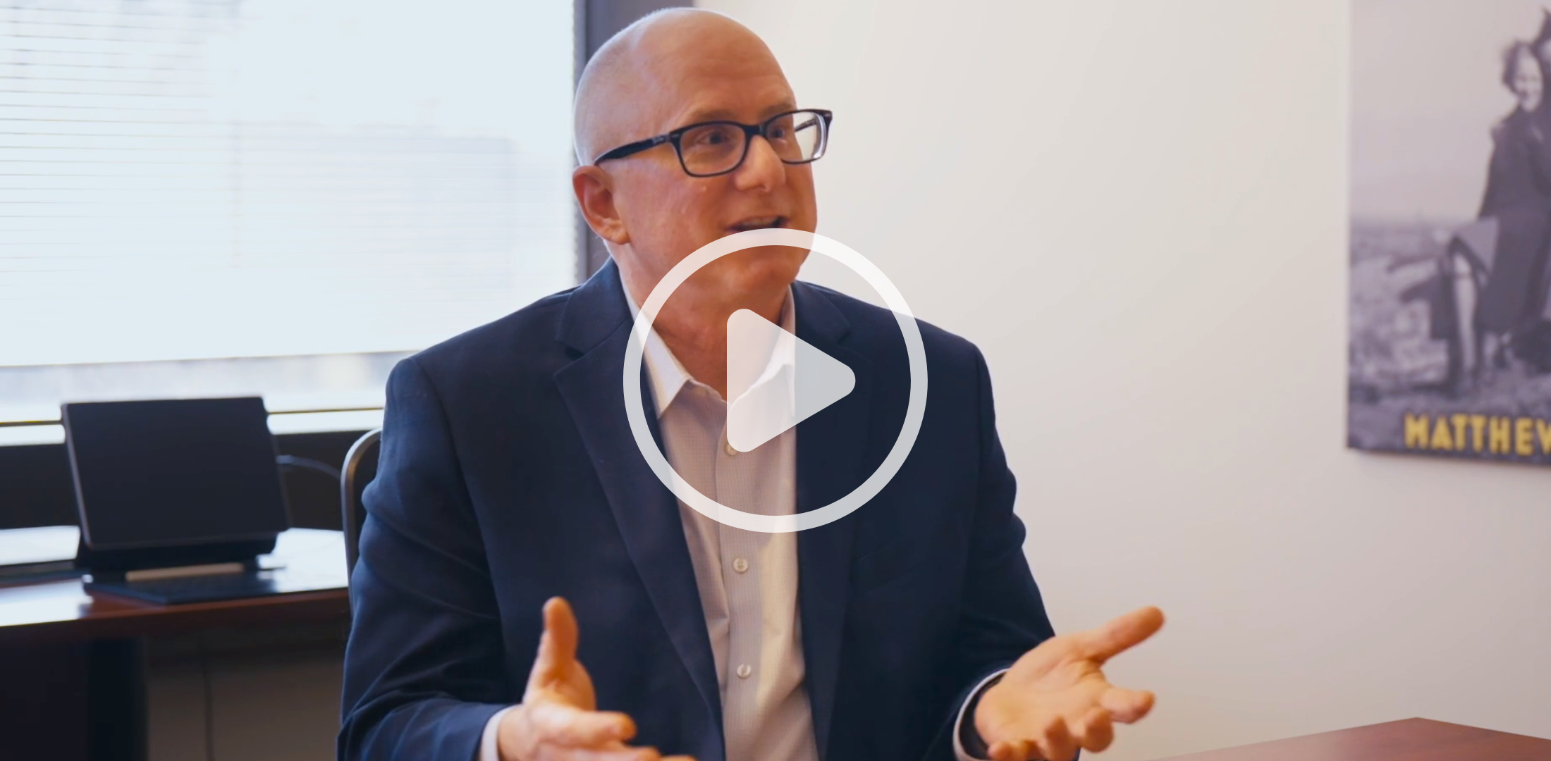 A man sits at a desk and is gesturing with his hands. There is a play button overlay on the image.