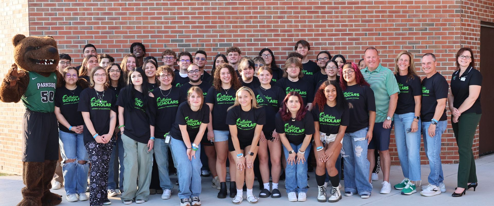A group of students, Callahan Scholars, smiling in front of the sports and activities center with the Callahan Family, Ranger Bear, Chancellor Lynn Akey, and Kenosha Mayor David Bogdala. 