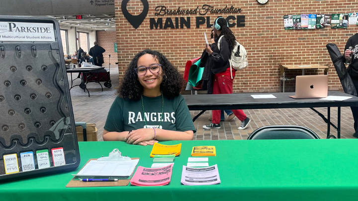 A student wearing a green Rangers t-shirt smiles behind a green-clothed information table at UW-Parkside