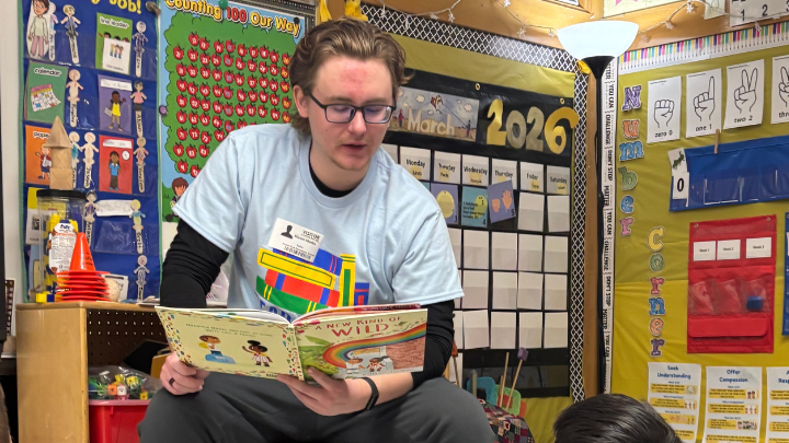 A student wearing a light blue t-shirt reads the children's book A New Kind of Wild to a classroom. The background is decorated with colorful educational posters, a March 2026 calendar, and student activity charts.