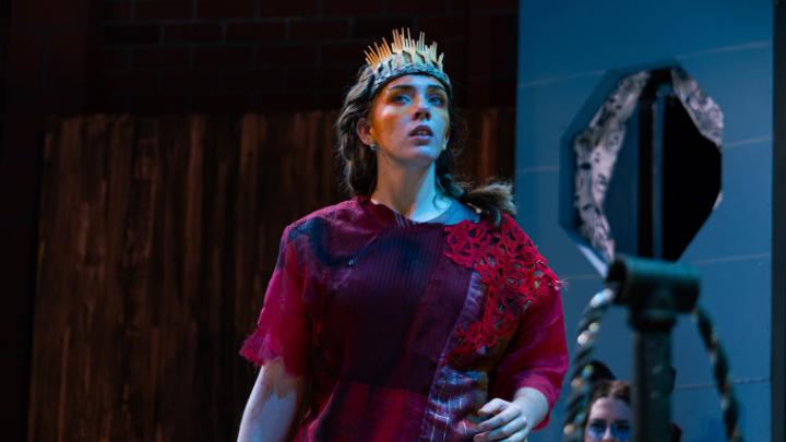 A student in a theatrical production looks upward with a dramatic expression. They are wearing a red, textured costume with lace detailing and a crown made of wooden spikes or tines. The background features dark, industrial-style stage elements.