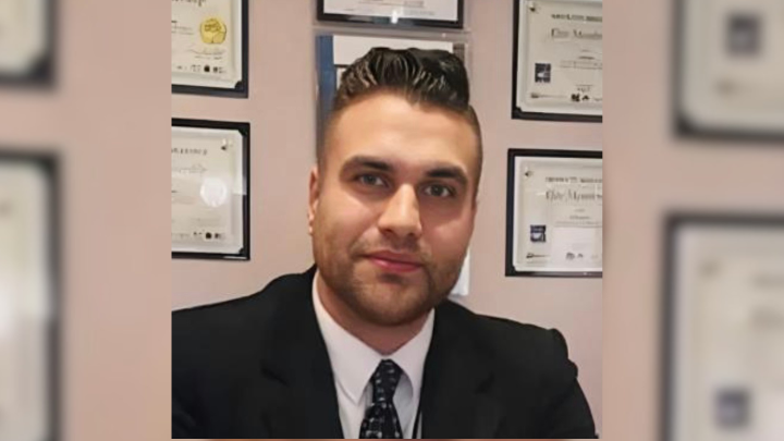 A professional portrait of a man with styled hair, a dark suit, and a patterned tie. He is looking toward the camera with a neutral expression. Several framed certificates or awards are visible on the wall behind him, creating a formal office-like setting