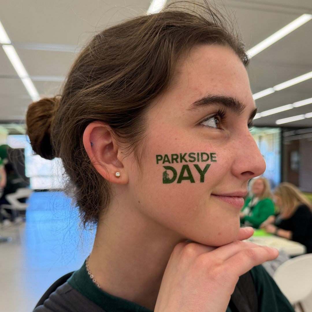  A close up profile shot of a person with brown hair in a bun looking off to the side. They have a green Parkside Day temporary tattoo on their cheek.