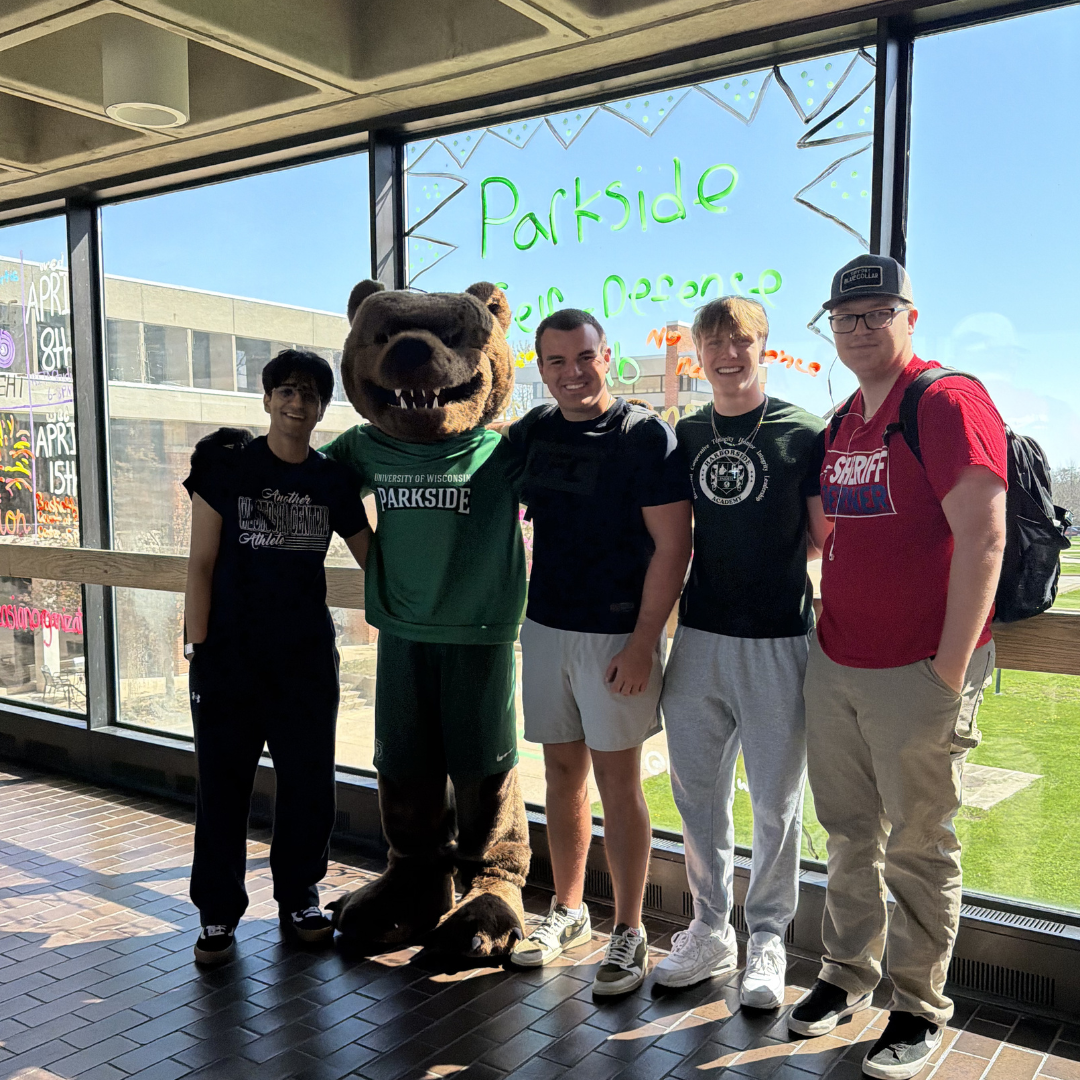  Four students stand together smiling alongside the UW-Parkside bear mascot in front of a large window on campus.