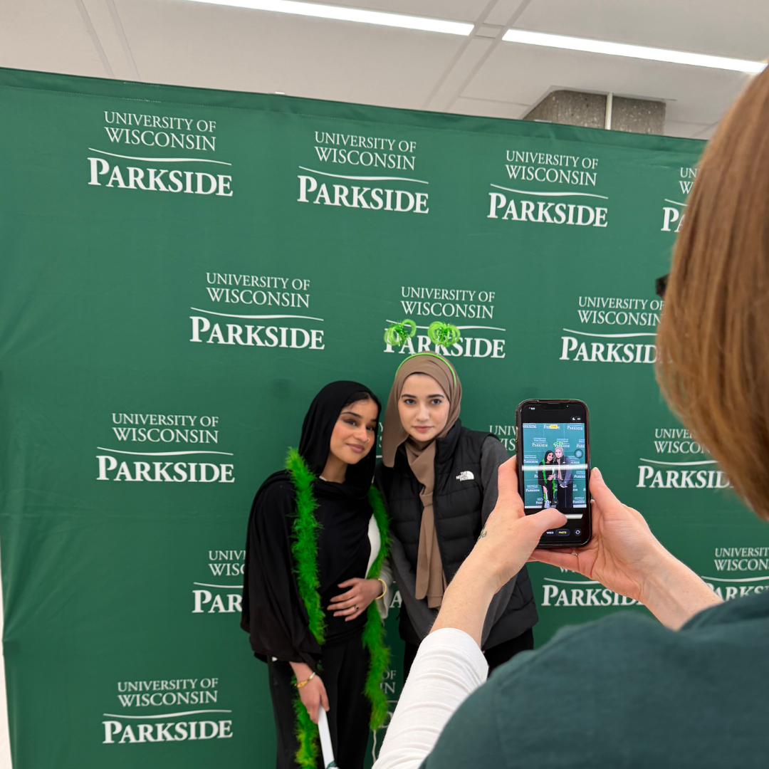  Two students pose for a photo in front of a green UW-Parkside backdrop. One student wears a green feather boa while the other wears festive green headband boppers as someone captures their picture on a smartphone.