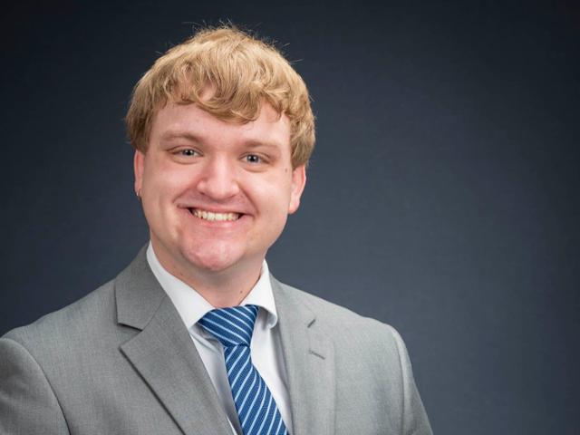 Photo of blonde man smiling in gray suit and blue tie. 