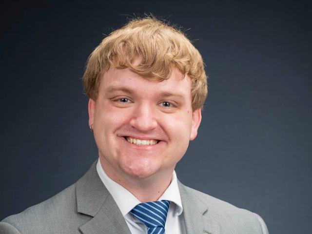 Photo of blonde man smiling in gray suit and blue tie. 