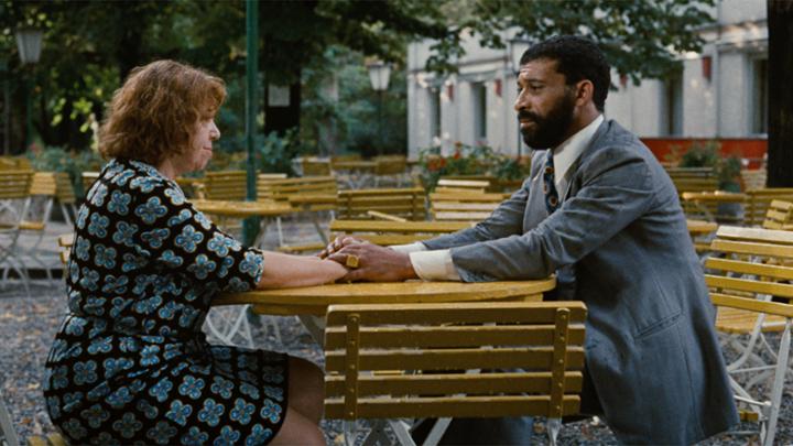 A woman and man sit across from each other at an outdoor table outside of a restaurant. He has his hands over hers on the table and they are looking at each other.