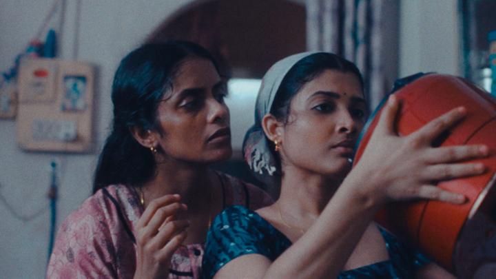 Two Indian women, one standing behind the other, looking at an orange rice cooker