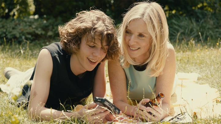 Two people laying in grass outside. Person on the left is showing something on a small device, the other person is holding a brown bottle.