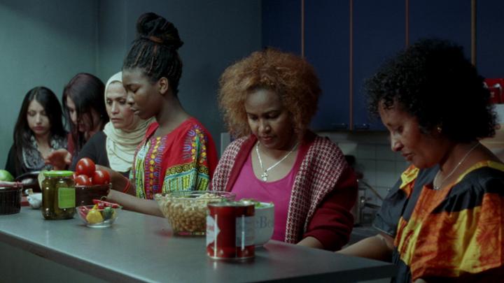 Six women sit at a kitchen counter with various vegetables looking down at their laps
