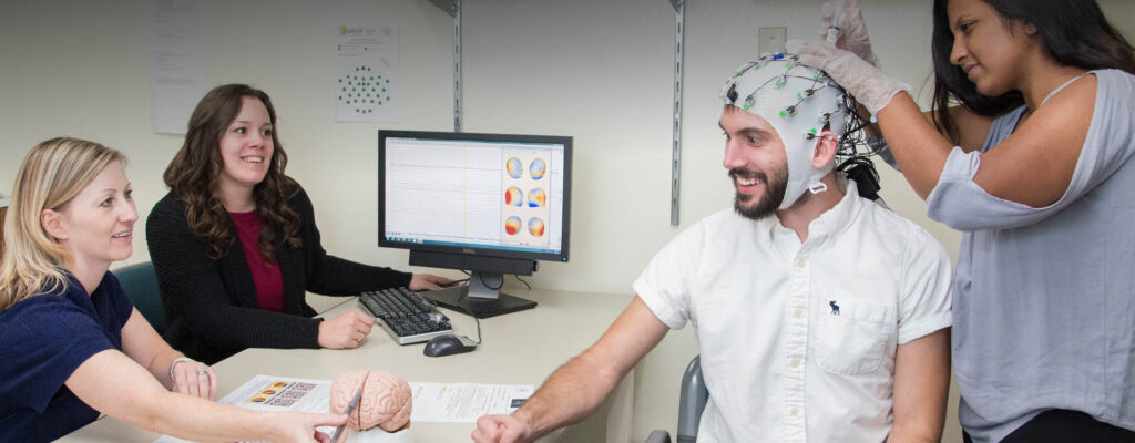 Four students doing psychology research. One student has a device connected to his head while another student helps attach it. To other students sit in front of a computer.