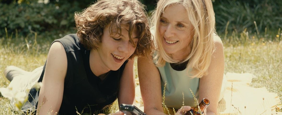Two people laying in grass outside. Person on the left is showing something on a small device, the other person is holding a brown bottle.