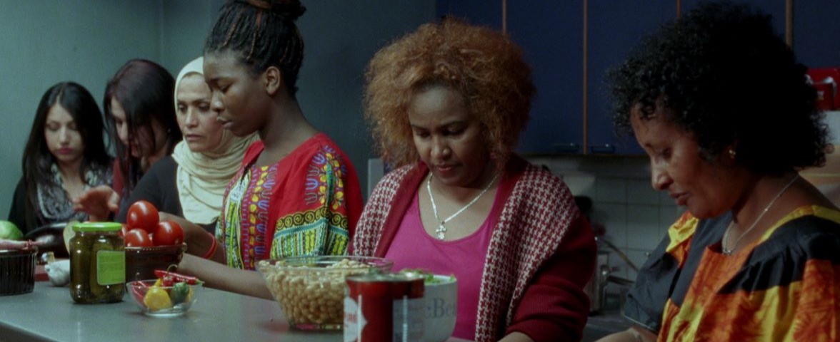 Six women sit at a kitchen counter with various vegetables looking down at their laps