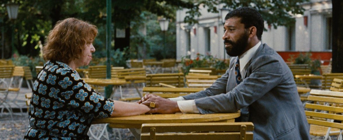 A woman and man sit across from each other at an outdoor table outside of a restaurant. He has his hands over hers on the table and they are looking at each other.