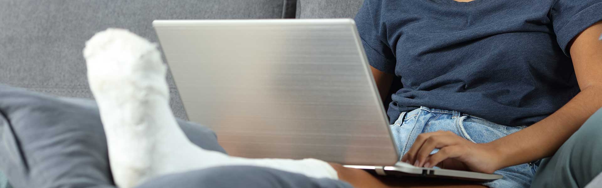 Person relaxing on a couch and working on a laptop, with one leg elevated in a white cast.