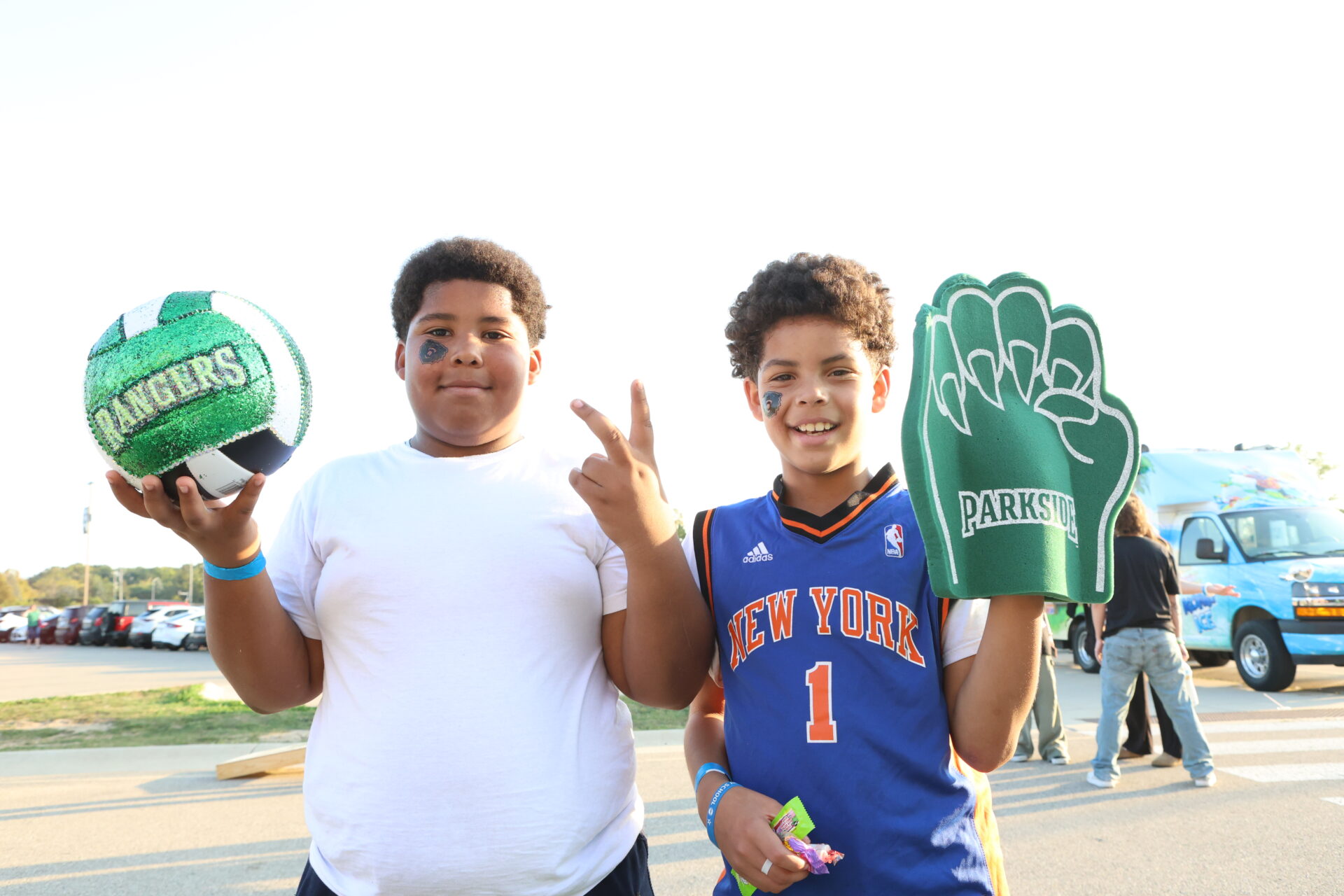 2 boys smiling at sunset in a parking lot. one is holding a volleyball and the other is holding a Ranger's Bear claw.
