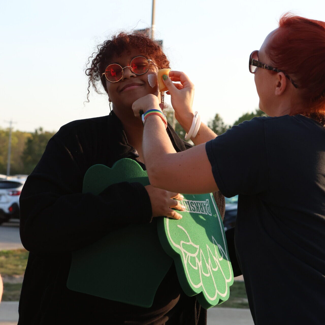 2 women in a parking lot at sunset. One woman is getting a temporary tattoo on her cheek, the other is applying the temporary tattoo.