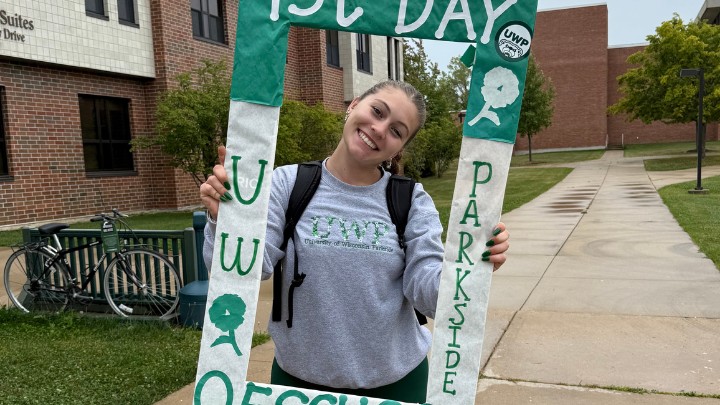 Gemini said A smiling young woman stands on a campus sidewalk, holding a large, handmade green and white photo frame that reads