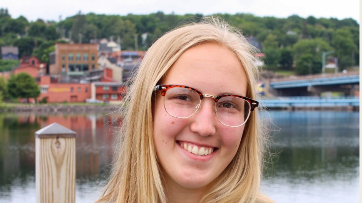 A close-up portrait of a young woman with long blonde hair and glasses, smiling at the camera. She is wearing tortoiseshell-rimmed glasses and a neutral-colored top.