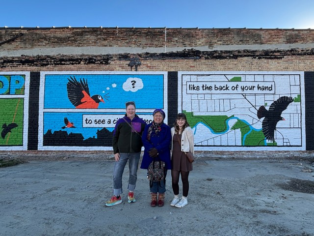 3 individuals stand in front of a colorful mural depicting birds flying above a map of Racine, Wisconsin